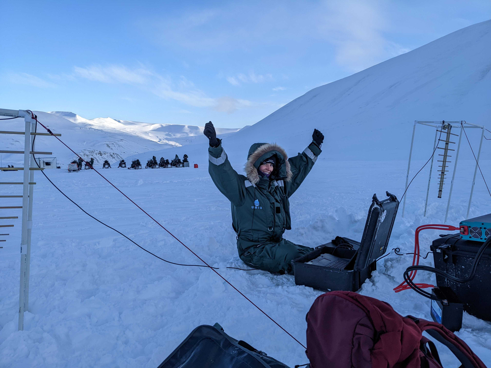 A young scientists lifts up their arms while sitting on the ice, located in Norway, while collecting data with an experimental ice-penetrating radar.