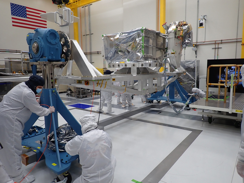 Two major pieces of flight hardware for the Europa Clipper spacecraft are attached to a flotron machine in a cleanroom. The hardware is visible in the rear middle of the images. The flotron is a large structure that allows the hardware to be rotated and moved for assembly purposes. The flight hardware consists of a large aluminum vault, which is shaped like a box. Within the vault, sensitive electronics will live, protecting them from radiation in Jupiter's environment. Attached the vault is a metal deck, called the nadir deck. Attached to this metal deck are multiple instrument sensors and cameras, which will allow the spacecraft to conduct science. Four engineers in full body coveralls and masks are seen working on flotron. An American flag is visible in the top left of the image. 