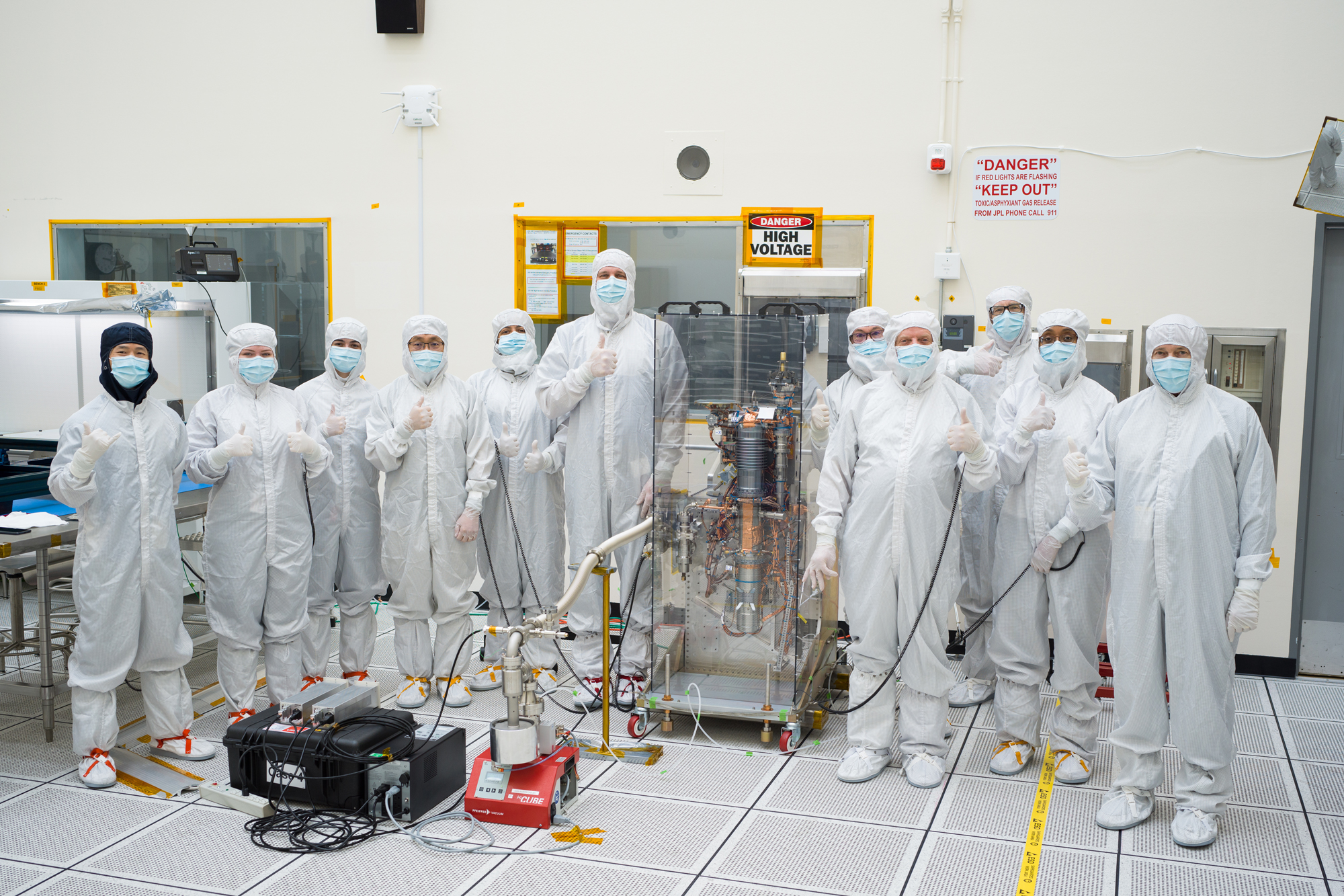 A group of eleven engineers wearing full-body white protective clothing stand facing the camera in a cleanroom. All of the engineers are giving a thumbs up hand expression. In the middle of the group of engineers is a large transparent box, approximately five feet tall, two feet wide, and two feet deep. Inside the transparent box is the mass spectrometer. Europa Clipper’s mass spectrometer takes up the middle of this image. The instrument consists of a long cylinder, several feet tall and a couple of inches in diameter. It takes the shape of an automobile spark plug, with a series of ridges visible at the top. Copper plating and wires extend from the instrument in multiple areas. The instrument is installed on a supportive structure.