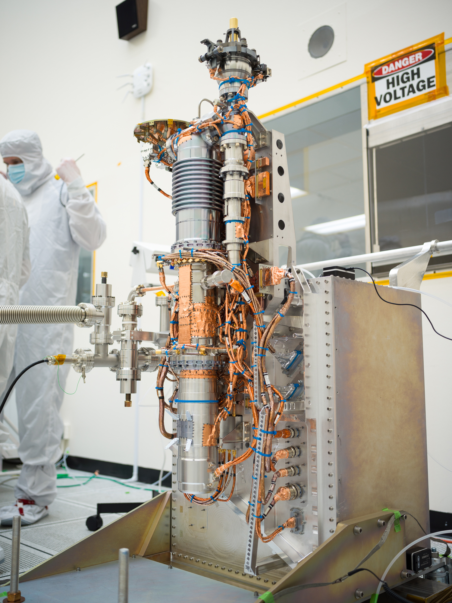 Europa Clipper’s mass spectrometer takes up the middle of this image. The instrument consists of a long cylinder, several feet tall and a couple of inches in diameter. It takes the shape of an automobile spark plug, with a series of ridges visible at the top. Copper plating and wires extend from the instrument in multiple areas. The instrument is installed on a supportive structure. Engineers are visible in the background working. 