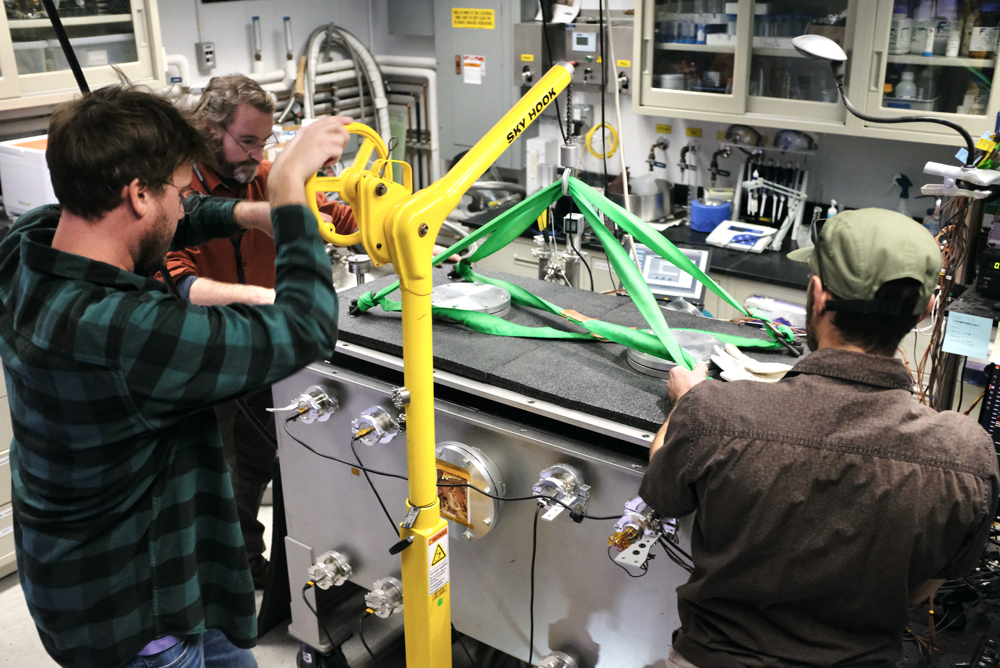 Inside JPL’s Ocean Worlds Lab, Jeff Foster, technologist, carefully cranks a bright yellow crane to lift the lid off of the Ark, a gray vacuum chamber about the size of a deep freezer. With Jeff are Dr. Kevin Peter Hand, Ocean Worlds Lab director, and Dan Berisford, JPL technologist. Both hold and guide green support straps connecting the crane to the Ark’s lid. Seen in the background are various tools, instruments, and cabinets.