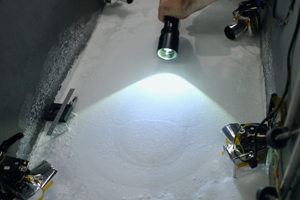  A hand holds a flashlight illuminating a large block of salty ice in a gray vacuum chamber about the size of a deep freezer. The top of the ice block features minor variations in texture, and is bordered by a collection of salts on the surface. Also shown are several small cameras mounted along the chamber’s interior walls, which are connected by wires stretching to the top of the image.