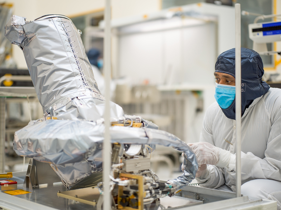 A worker kneels beside the Europa Clipper’s narrow-angle camera (NAC), which is on a metal platform with rods sticking straight up at the corners of the platform. The worker is covered head-to-toe in white protective clothing called a bunny suit. The worker is wearing a blue head covering, and a bright blue face mask. The NAC is wrapped in silver-colored insulating material, it has a round base about the size of tire. The lens is sticking out at an angle and is laced up inside the insulating material. 