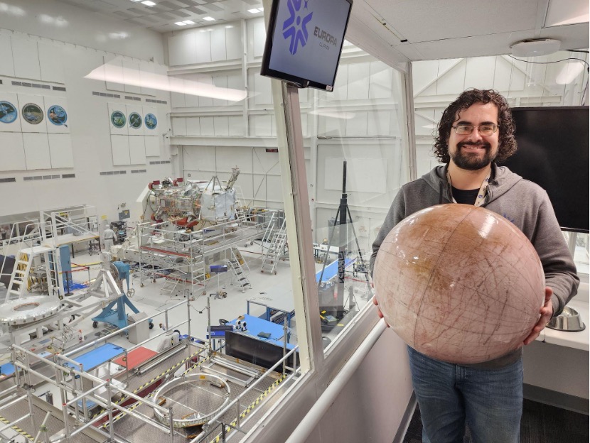 Stephen Pakbaz, JPL mechanical engineer, holds a beach ball-sized model of Europa overlooking the High Bay 1 Viewing Gallery at NASA’s Jet Propulsion Laboratory (JPL) in Southern California. Seen in the background behind Stephen are groups of engineers assembling components of the Europa Clipper spacecraft.