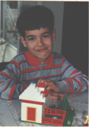 Shown in this photo is Stephen Pakbaz, JPL mechanical engineer, from his youth. Stephen sits at a table and proudly displays a toy model house made out of red and white interlocking brick pieces with a green tree on an adjacent front yard.