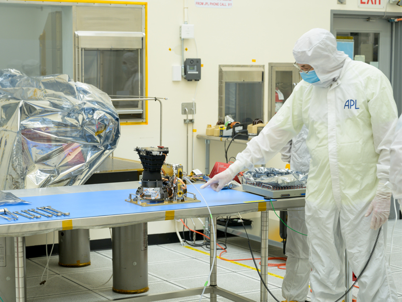 An engineer in full white coveralls and a mask stands pointing at Europa Clipper's wide-angle camera, while inspecting the instrument in a clean room. The wide-angle camera rests on a silver metallic table in a clean room. 