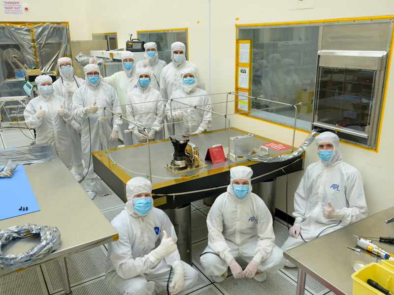 Eleven engineers wearing full white coveralls and masks stand around a metallic table in a clean room. Europa Clipper's wide-angle camera rests on the metallic table. Several engineers are giving a thumbs up hand gesture, indicating that the instrument has successfully undergone functional tests following its delivery to JPL.  