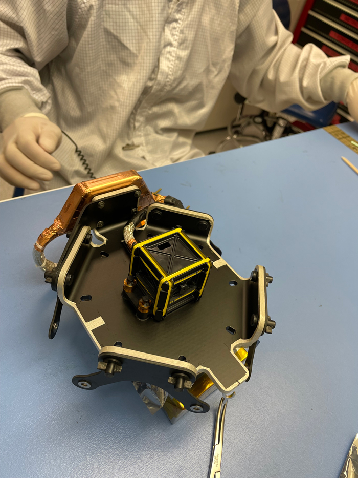 An octagonal device about the size of a small toaster sits on a laboratory table before a worker wearing a white full-body covering in a clean room at NASA's Jet Propulsion Laboratory.