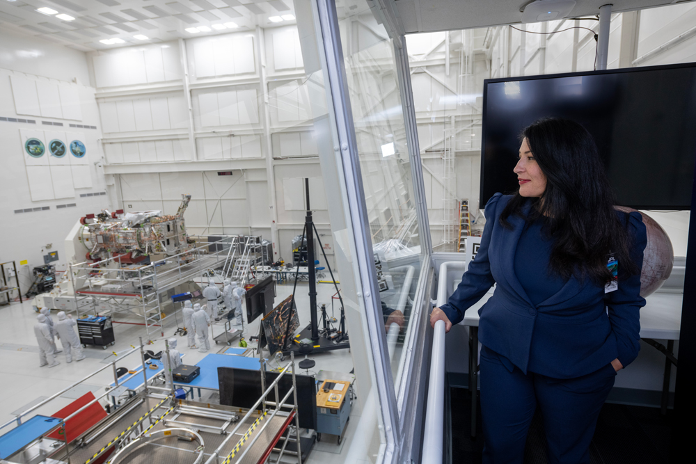 U.S. Poet Laureate Ada Limón is standing on the right side of this image, wearing a dark blue suit and looking to the left. A glass wall separates Limón from the clean room, which is visible on the left side of the image. In the distance, the Europa Clipper is visible as it is being assembled in the clean room. It is in a horizontal position with scaffolding surrounding it. Approximately nine engineers are visible near the spacecraft wearing white full-body coveralls.