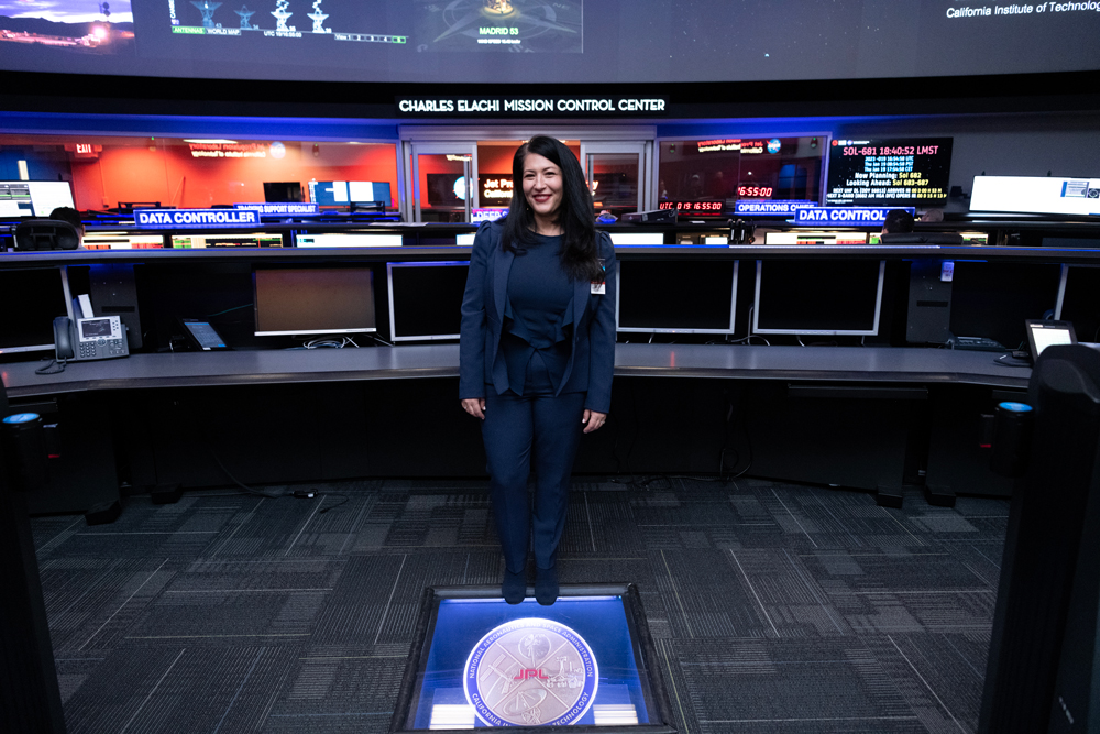 U.S. Poet Laureate Ada Limón is standing in the center of this image, facing the camera, wearing a dark blue suit. Behind Limón is a long console stretching from both sides of the image with multiple screens. Signs in the background read “Data Controller”, and a sign on the wall behind Limón reads “Charles Elachi Mission Control”. Limón is standing in front of a glass light box that is inlaid into the carpeted floor. Within the box is a metal plaque that reads “Center of the Universe”. 