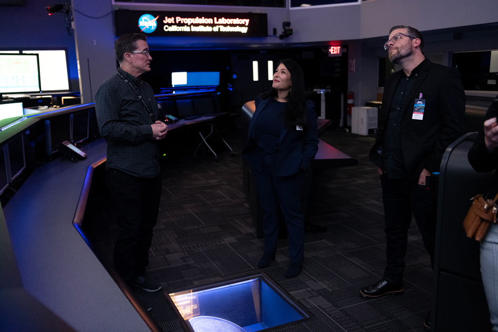 U.S. Poet Laureate Ada Limón is standing in the center of this image, looking up. She is surrounded by consoles with screens and a sign on the back reads “Jet Propulsion Laboratory, California Institute of Technology” next to the NASA meatball logo.