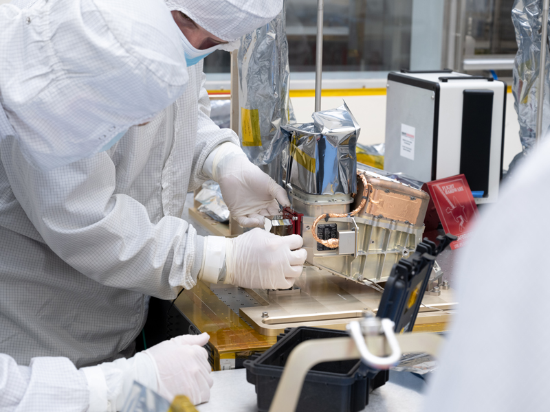 Multiple engineers in white coveralls preparing the thermal imager instrument, located on a table in a clean room, for functional testing. One engineer has their hands on the instrument. 