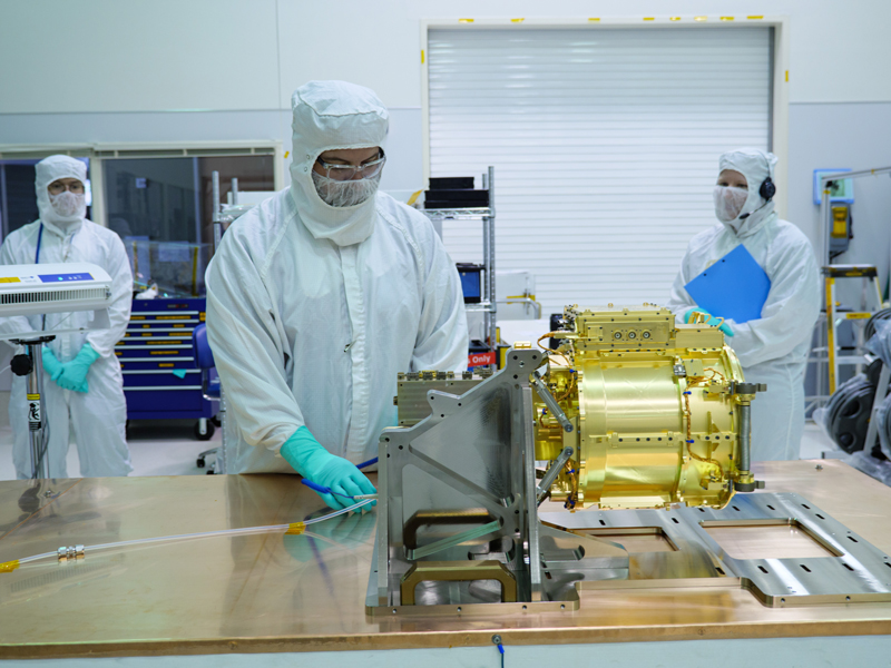 The SUDA sensor head rests upon a table in a clean room. The sensor head is gold in color, shaped slightly like a drum on its side. It is attached a device that lifts the sensor head from the table, and is seen slightly to the ride of center in the image. An engineer wearing white full body coveralls, light blue gloves, glasses, and a mask stands behind the sensor head and the table it is on, looking down at the sensor head. Two additional engineers stand in the distance on either side of the sensor head, both wearing full white body coveralls, masks, and light blue gloves.  