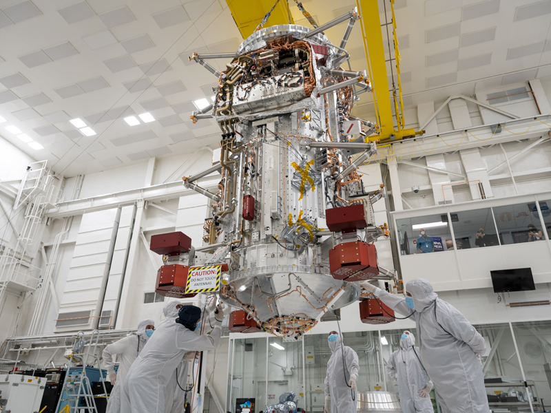 In the center of the image, the main body of the Europa Clipper spacecraft hangs from a yellow crane in a large, multistoried cleanroom with white walls and a white ceiling. Six engineers in full body coveralls and masks stand around the base of the main body as it hangs from the crane, with some working on the main body. In the distance, a viewing gallery is visible with four people watching the activities taking place. 