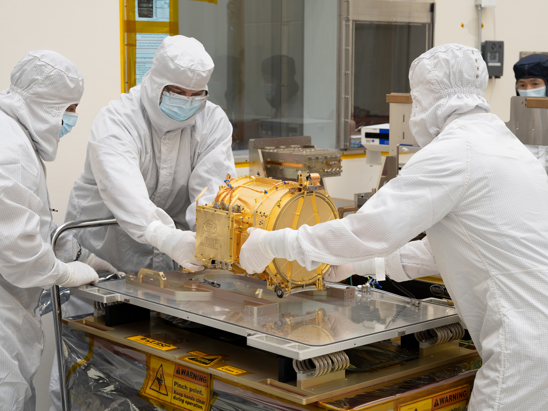 Two engineers in full white body coveralls are seen lowering Europa Clipper’s surface dust analyzer onto a silver workbench in a cleanroom. Another engineer standing to the left, with a hand resting on the silver workbench. A fourth engineer is seen observing in the distance. The instrument is gold colored and about the size of a drum, resting on its side. The instrument is resting upon a silver workbench in the cleanroom. 