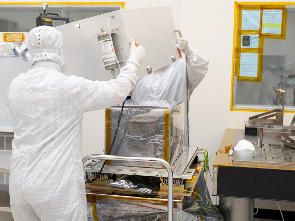 Two engineers in full white body coveralls and masks are shown in the center of the image lifting a silver box up from a cart, revealing Europa Clipper’s surface dust analyzer instrument underneath. The engineers and the instrument are seen in a cleanroom. The instrument is surrounded by a semi-transparent protective plastic that is in the shape of the box that was covering it. The gold colored instrument is about the size of a drum. 