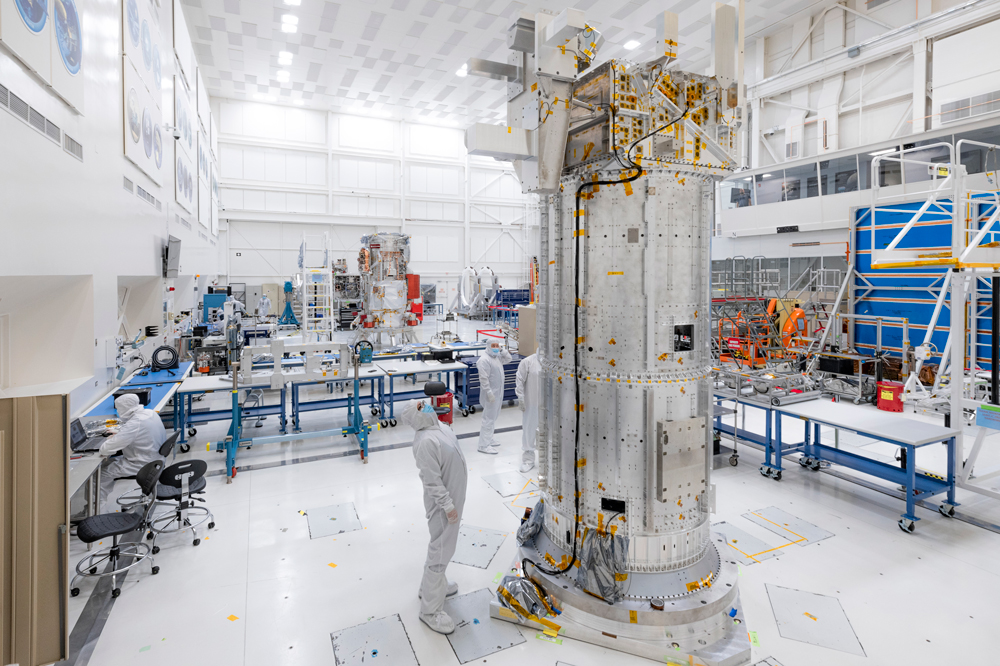 In the center of the photo, an engineer stands to the left of Europa Clipper’s developmental test model, which is visible as a large, aluminum cylinder with an aluminum box and other aluminum structures protruding from the box. The aluminum box replicates the spacecraft’s avionics vault, while the cylinder replicates the spacecraft’s propulsion module. The test model is towering more than twice as high as the engineer, who is wearing full white body scrubs. To the left and further back in the clean room, the spacecraft’s flight model is visible. 