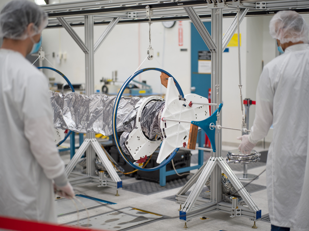 Two workers in white lab coats, cleanroom bouffant caps, and blue face masks have their backs to the camera as they unfurl as Europa Clipper’s magnetometer boom. A blue frame that holds the boom can be seen. The boom is supported by several silver-colored stands.