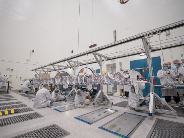 Several workers in white lab coats, cleanroom bouffant caps, and blue face masks work around Europa Clipper’s magnetometer boom after the boom was unfurled. Some of the workers are on the floor beneath the boom. Others are standing nearby. The boom is covered in protective silver-colored material and is supported by several aluminum-colored stands. 