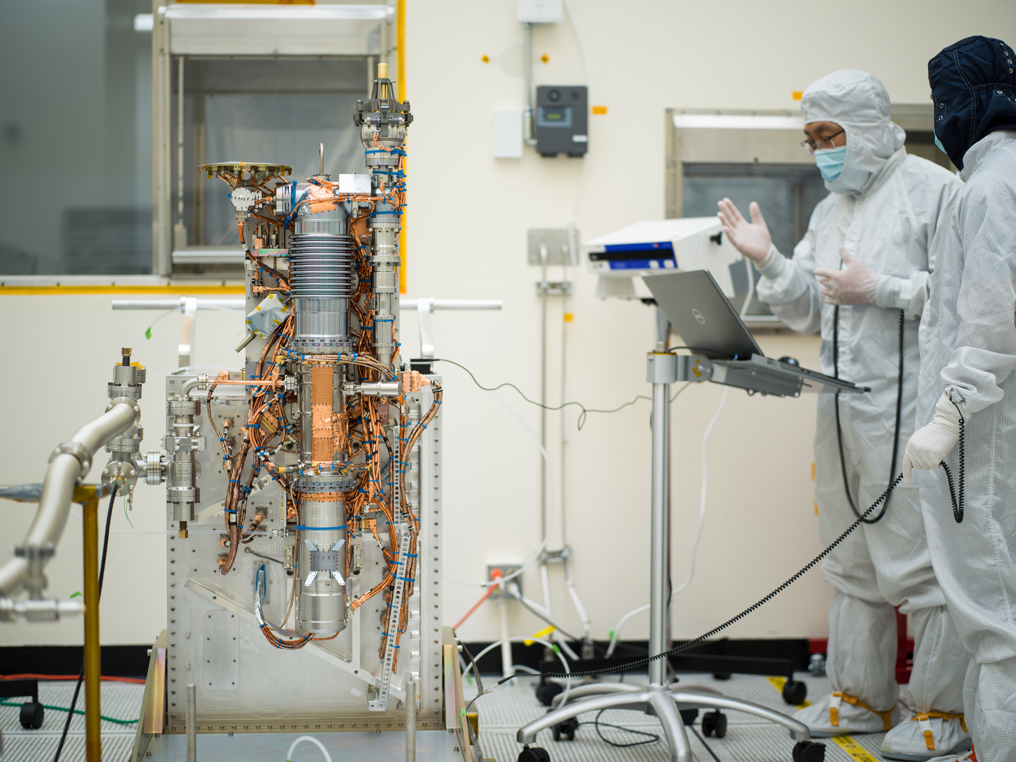 Europa Clipper’s mass spectrometer takes up the left side of this image. The instrument consists of a long cylinder, several feet tall and a couple of inches in diameter. It takes the shape of an automobile spark plug, with a series of ridges visible at the top. Copper plating and wires extend from the instrument in multiple areas. The instrument is installed on a supportive structure. Two engineers are visible on the right side of this image as they look at a laptop while performing tests on the instrument. 