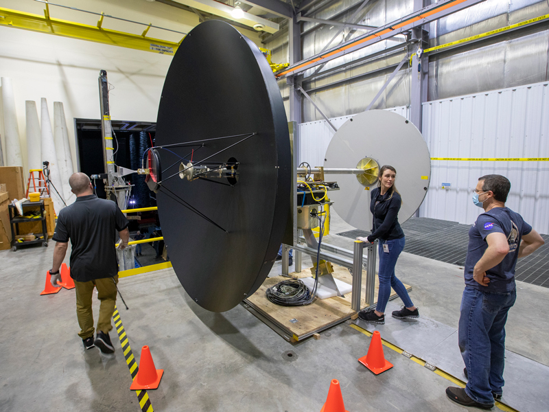 Researchers stand next to Europa Clipper’s high gain antenna, which is taller than a person and is painted dark black, as they prepare to move it into a testing facility. 