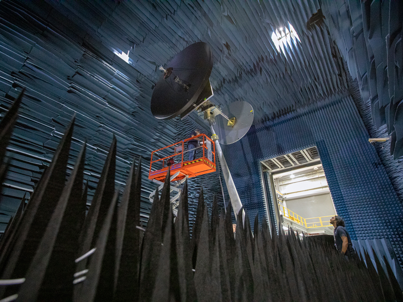 Europa Clipper's large high gain antenna, which is taller than a person and is painted dark black, is hoisted on mechanical supports on a large, multi-story room that has its floor, walls, and ceiling covered in blue foam triangles to avoid interference during testing. 