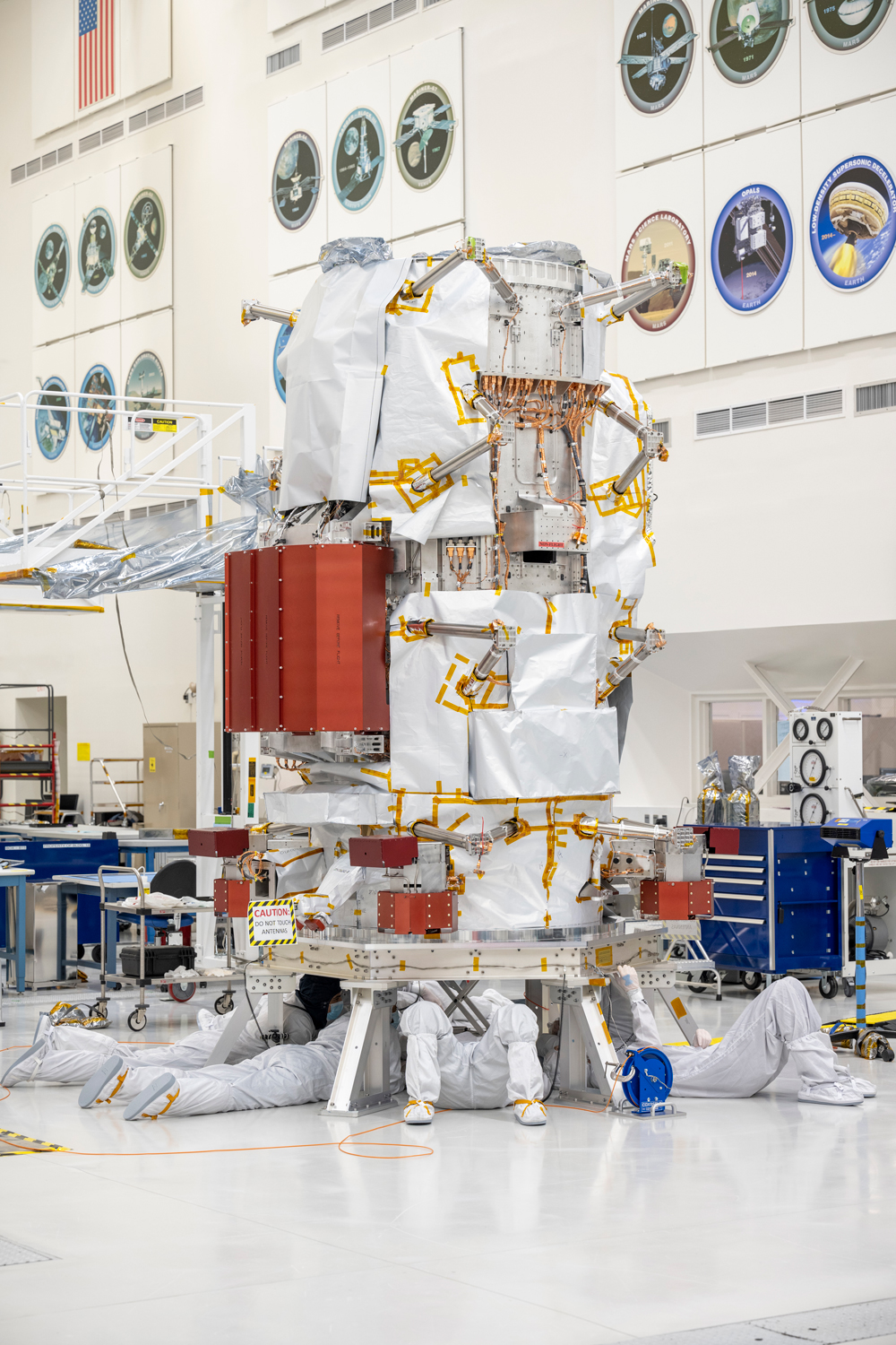 The main body of the spacecraft standing in a white clean room is the central component of this image. The tall structure is covered with protective covers to keep sensitive equipment safe during assembly and what looks like white paper, which is being used for patterning for the spacecraft’s protective insulation. Four engineers are seen under the base of the spacecraft, where they are installing the reaction wheels, as if they are working on the undercarriage of a car. Two engineers on the left are on their stomachs, and two engineers on the right are on their back. 