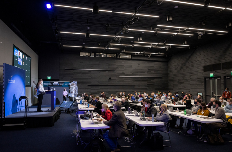A woman stands on stage on the left addressing a group of team scientists on the right, who are seated at multiple tables. 