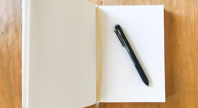 A blank notebook with white papers is visible open on a wooden table, with a pen laid over it. 