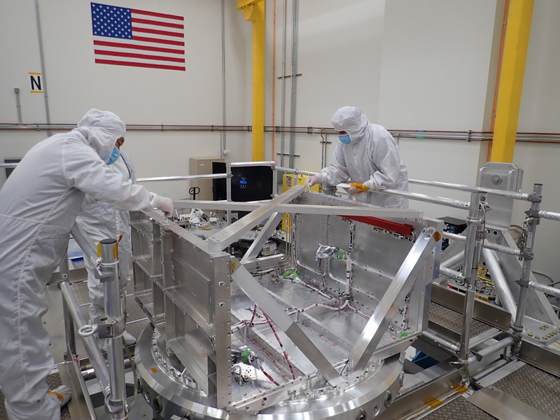 Three engineers wearing full-body coveralls, gloves, and masks use special tools to wipe fastener holes along the top edges of a hollow metal box about the size of a large living-room chair.