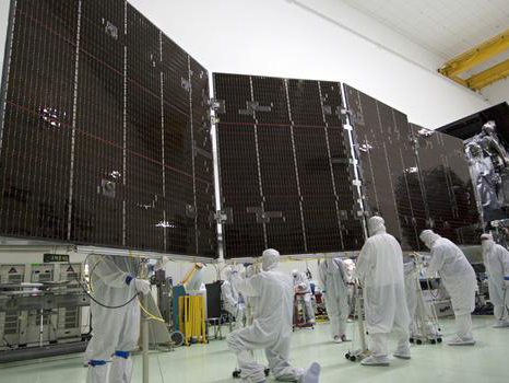 Large solar panels for NASA's Juno spacecraft are visible in a clean room being deployed, surrounded by engineers in full body coveralls. 