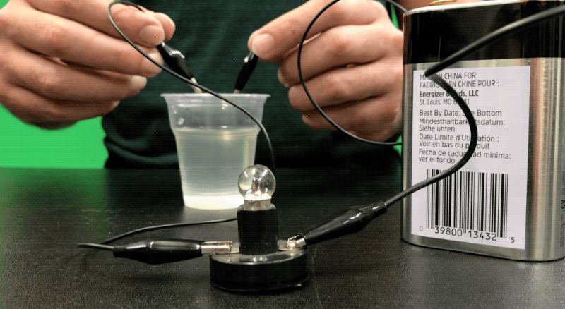 A person is seen holding two wires, one in each hand, and they are putting the wires into a clear plastic cup filled with water. 