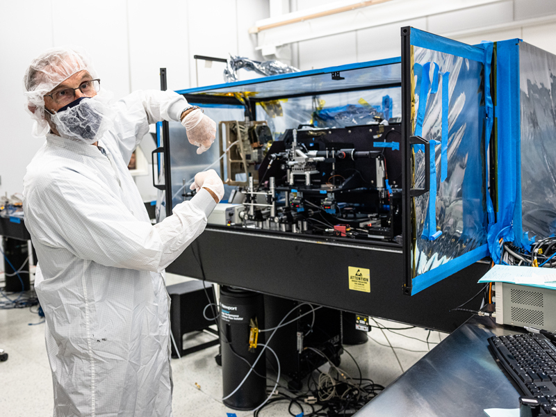 Instrument designer Phillip Christensen stands in white coveralls and a mask next to a plastic box tent structure, with the thermal imager and optical test equipment located inside. 