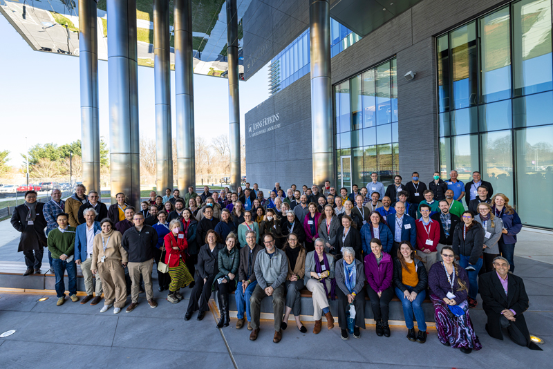 A large group of Europa Clipper science team members seated and standing on the steps of the entrance of a building at the Johns Hopkins University Applied Physics Laboratory’s South Campus. 