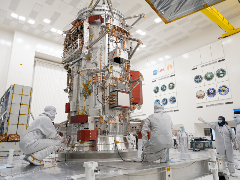 In a large white multistory room, the main body of the Europa Clipper spacecraft rests upon a stand as it takes its position in the ultra-hygienic High Bay 1 in the JPL's Spacecraft Assembly Facility. The main body is composed of a shiny metallic cylinder with wiring and electronics attached to it. Two people in full white coveralls and masks crouch next to the main body. A person in full coveralls and a mask stands in the distance watching. On the right, two additional people in coveralls and masks watch the work being done on the main body of the spacecraft, with one pointing at the main body. In the distance, the cleanroom's wall of fame is visible, with the identifiers of previous missions that have been assembled in the cleanroom. 