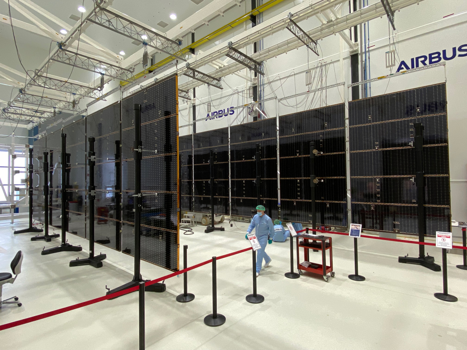 The spacecraft’s large solar array wings take up the majority of this image. They are parallel to each other in position and the image is taken at angle, so you can see the solar arrays extended into the background of the image. Each solar array wing consists of five panels connected to each other to form a long solar array wing that is approximately 46.5 feet (14.2 meters) long. Bronze colored lines are visible creating a grid pattern on each panel. Each solar array wing is hoisted several feet above the ground by support structures that attach to each solar array wing panel. The solar array wings are tall with an approximate height of 13.5 feet (4.1 meters). Walking between the solar array wings is an engineer wearing blue protective clothing. The engineer doesn’t even appear to come up to half the height of the solar array wings. The solar array wings are visible in a large white cleanroom, and the area with the wings is cordoned off with a red barrier. 