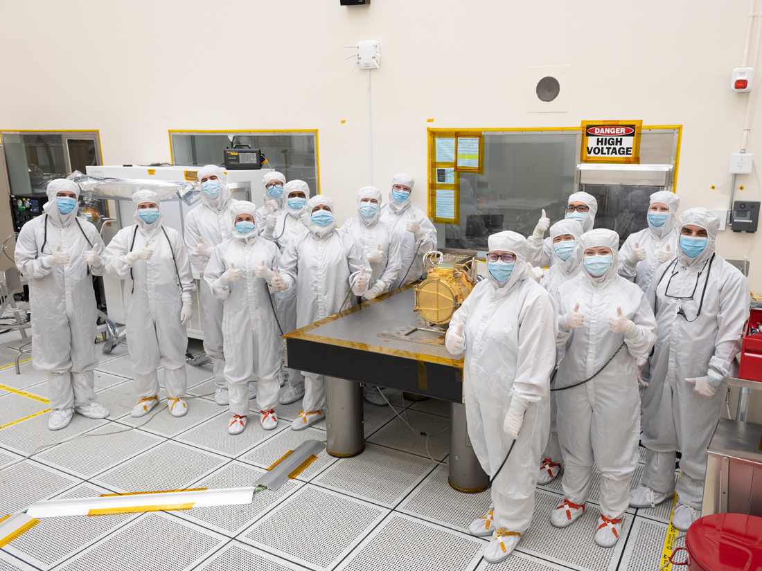 A large group of engineers in full white body coveralls stand surrounding Europa Clipper’s surface dust analyzer. All the engineers are giving a “thumbs up” hand gesture to indicate that the instrument was successfully delivered. Nine engineers stand to the left of a silver workbench, on which the instrument rests, and six stand to the right of the workbench.  The instrument is gold colored and about the size of a drum, resting on its side. The instrument is resting upon a silver workbench in the cleanroom. 