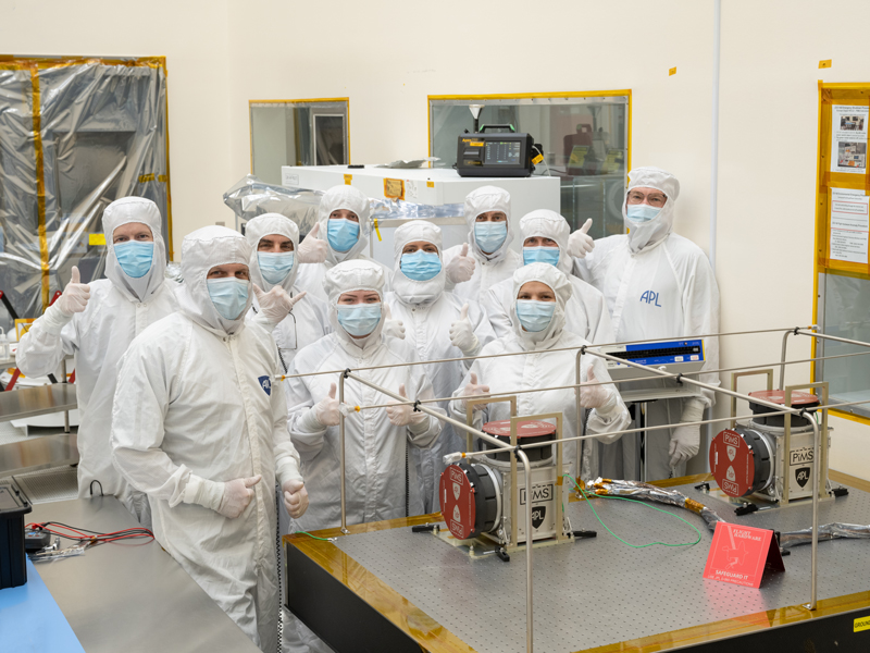 A group of engineers in full white coveralls with masks stand in a clean room. Several engineers are giving a thumbs up hand gesture. They are standing around two faraday cups, part of the spacecraft's plasma detection instrument, following functional tests performed after the instrument was delivered to NASA's Jet Propulsion Laboratory in Southern, California.