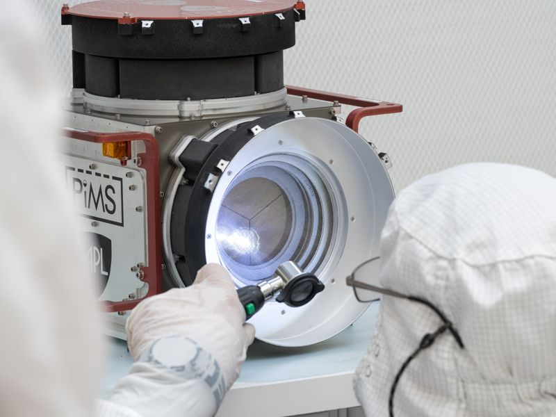 An engineer examines a faraday cup, part of Europa Clipper's plasma detection instrument, which is resting on a table in a clean room. The engineer is using a flash light to examine the instrument. 