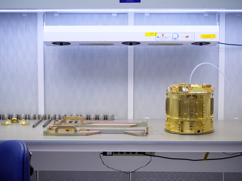 The SUDA sensor head is shown resting on a table in a clean room, seen on the right-hand side of the image. The sensor head is gold in color and looks slightly like a drum in shape. A clear tube is seen attached to the top, where nitrogen gas is pumped through. To the left of the sensor head, additional equipment is shown resting on the table. Above the table a fan device is shown hanging from the ceiling.  