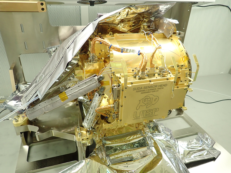 The instrument sensor head is shown in the middle of the image as a large metallic device, gold in color, with wiring attached. The sensor is resting on a table in a cleanroom. There are no people visible in the image. Multi-layer insulation is visible on the top and underneath the instrument. On the side of the instrument is a panel with information etched in, including “SUDA SENSOR HEAD”, “LASP” (which refers to the Laboratory for Atmospheric and Space Physics), and the mascot symbol for the University of Boulder, which is a buffalo. 