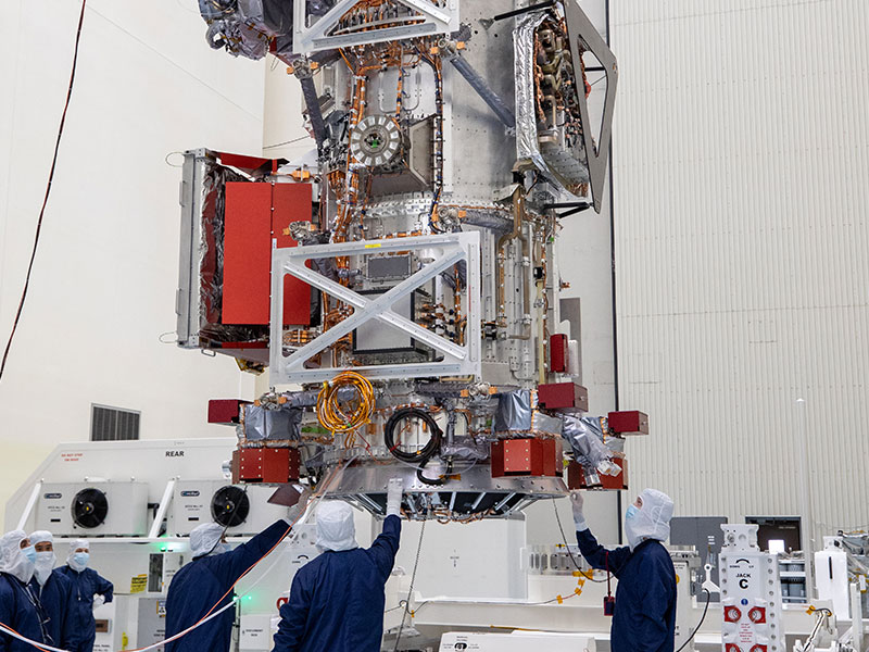 Workers in blue overalls and white head covers help guide Europa Clipper as a crane hoists it as part of prelaunch processing.