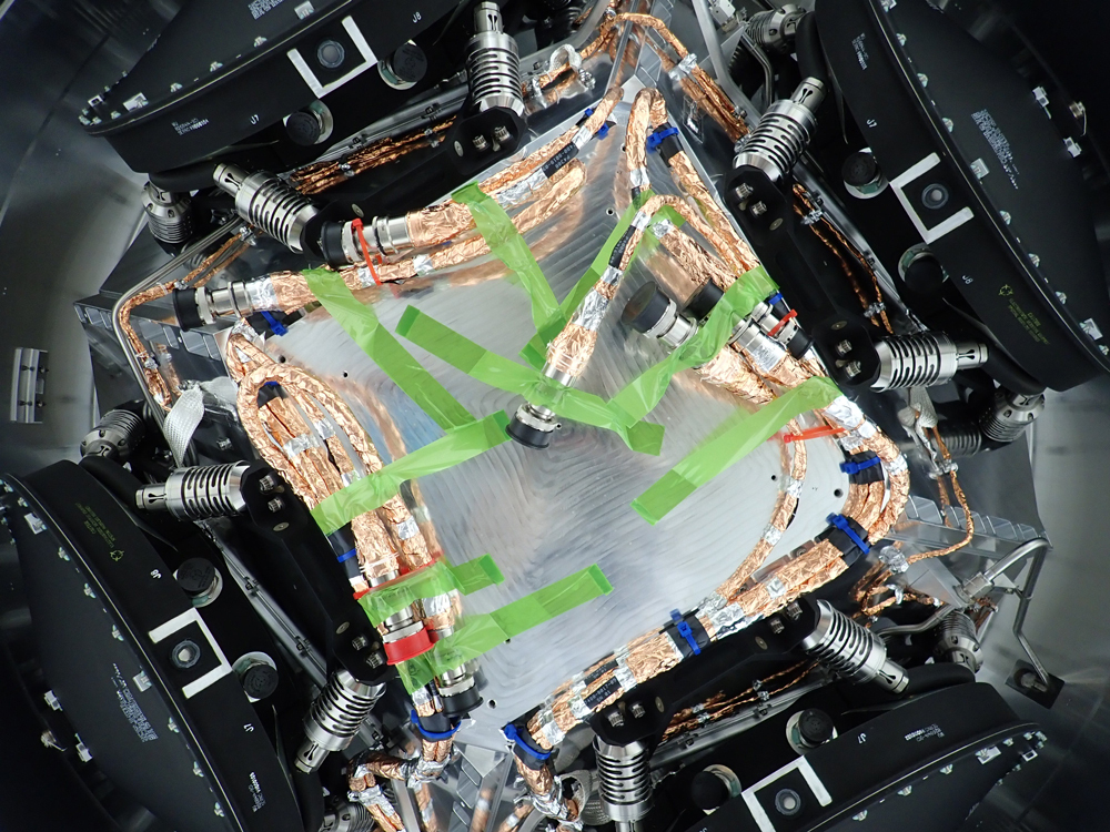 Four reaction wheels are seen here, one in each of the four corners of the image. The reaction wheels are black. In the configuration shown, the wheels are visible from their sides, which allows you to see the wiring and other mechanical equipment attached to the reaction wheels. In the middle of this image you can see a silver triangle, which is base of the main body of the spacecraft. Thick bundles of gold wires extend from the reaction wheels, with green tape holding the bundles into place as the spacecraft continues through assembly. 