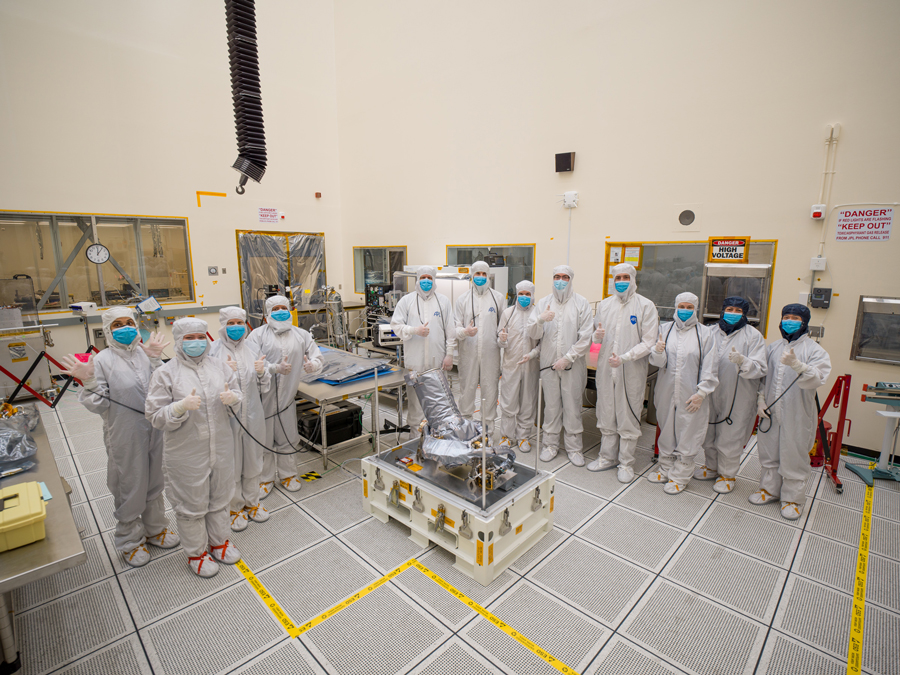 Twelve engineers and technicians dressed in white protective “bunny suits” stand behind and beside to Europa Clipper’s narrow-angle camera (NAC). The NAC is on a platform with rods sticking straight up at the corners of the platform. It’s wrapped in silver-colored insulating material, it has a round base about the size of a tire. The lens is sticking out at an angle and is laced up inside the insulating material. Yellow tape on the floor marks the area around NAC. Equipment and gear sits on tables behind, and to the sides of the workers. 