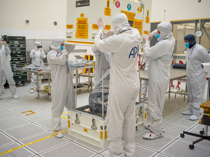 In the center of the image, four technicians are seen lifting off a square, white protective cover from a white container that holds Europa Clipper’s narrow-angle camera. The engineers are wearing white full-body coveralls, including blue face masks. Two are facing away from the camera and two are facing the camera. The two closest to the camera have APL written on the back of the coveralls, which stands for “Applied Physics Lab”. Another worker in a white bunny suit and dark blue hood stands off to the right side of the image. A wall clock is to the right of his head. To the far left of the image, two people in bunny suits are recording the moment: One person is holding a video camera, and the other is holding a smartphone. A toolbox is against a wall on the left side of the room.