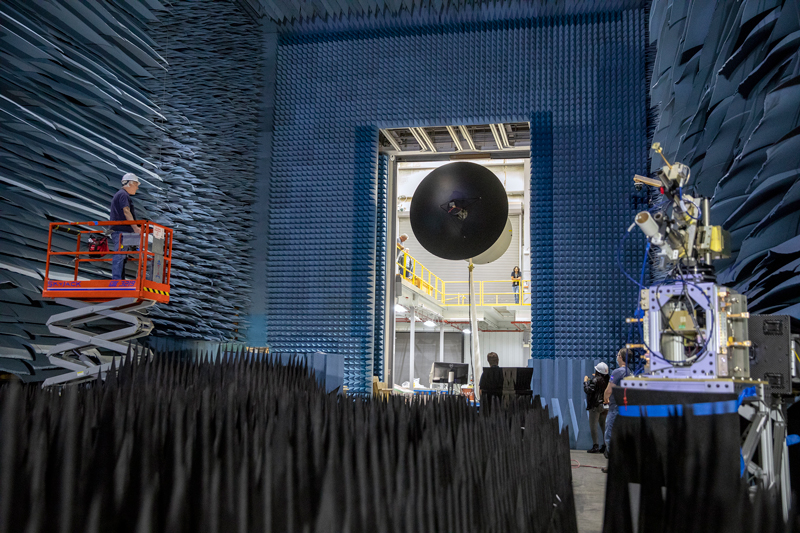 Europa Clipper's large high gain antenna, which is taller than a person and is painted dark black, is hoisted on mechanical supports on a large, multi-story room that has its floor, walls, and ceiling covered in blue foam triangles to avoid interference during testing. 