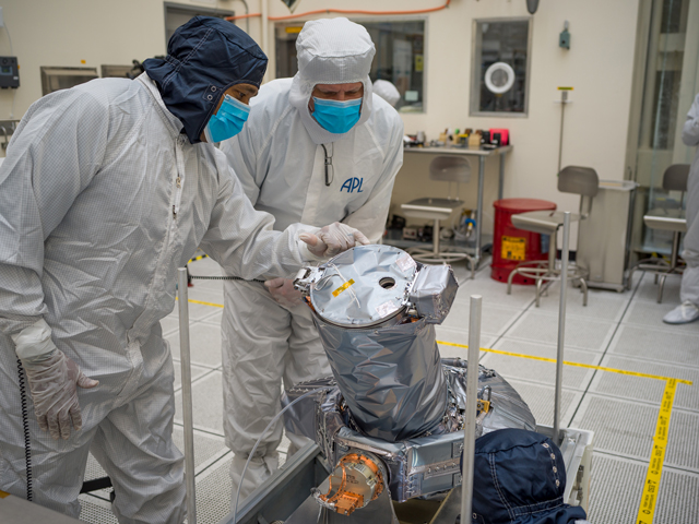 Two workers are standing beside the Europa Clipper’s narrow-angle camera (NAC), which is on a metal platform with rods sticking straight up at the corners of the platform. The workers are covered head-to-toe in mostly white protective clothing called bunny suits, and they are wearing bright blue face masks. One worker is leaning toward the NAC and pointing to the NAC, which is wrapped in silver-colored insulating material. The NAC has a round base about the size of tire. The lens is sticking out at an angle.