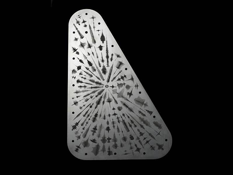 An image of the Europa Clipper vault plate, highlighted from the upper left corner casting shadows that emphasize engraved waveforms emanating from a central point symbolizing the word 'water' in multiple languages. The waveforms present as audio frequency waves. The vault plate is metal silver and the engravings are black, set against a black background. 
