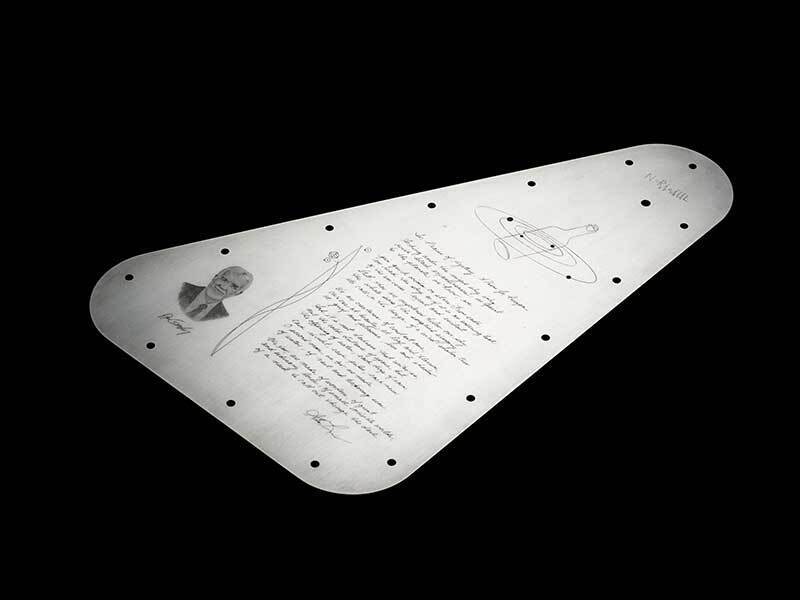 A full view of the Europa Clipper vault plate’s inward-facing side. The upper portion features the Jovian system with a bottle replacing Jupiter, and the lower portion is dominated by a handwritten poem in elegant script. A lifelike engraved portrait sits to the left of the poem. The plate has a symmetrical arrangement of holes along its perimeter for mounting. The background is black, highlighting the metallic sheen of the plate and the details of the engravings.