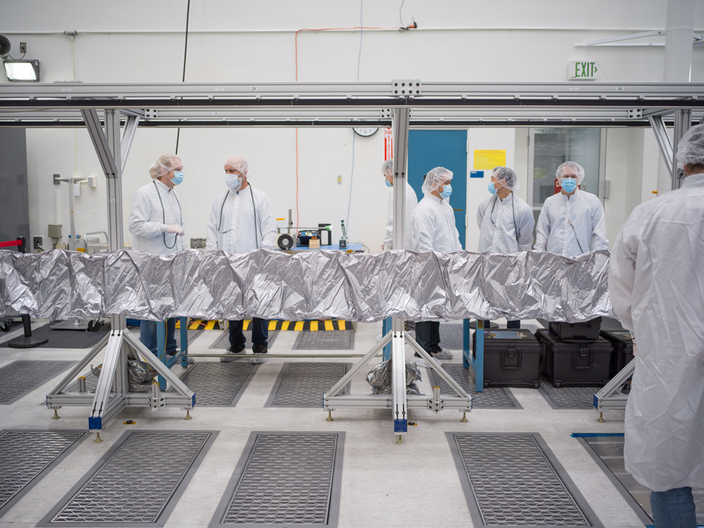 Seven workers in white lab coats, cleanroom bouffant caps, and blue face masks stand near Europa Clipper’s magnetometer boom after the boom was unfurled. The boom is covered in protective silver-colored material and is supported by several aluminum-colored stands. 